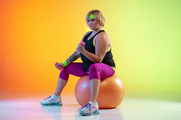 Young caucasian plus size female model's training on gradient orange background in neon light.