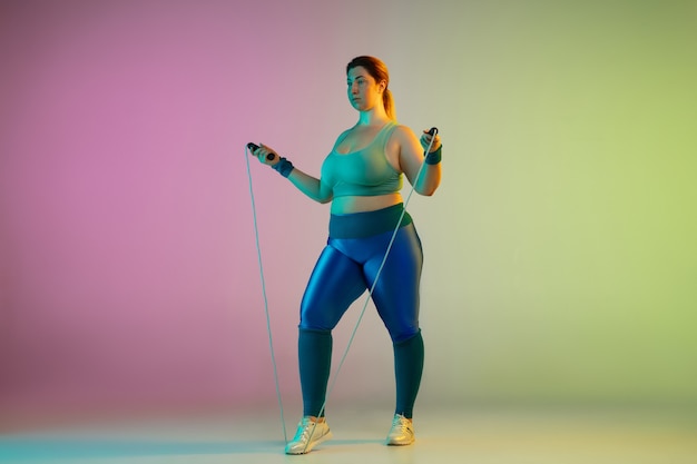 Young caucasian plus size female model's training on gradient purple green wall in neon. Doing workout exercises with jump rope.