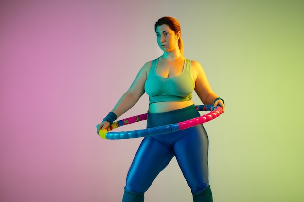 ネオンの光の中でグラデーション紫緑の壁に若い白人プラスサイズの女性モデルのトレーニング。フープでトレーニングエクササイズをする。スポーツ、健康的なライフスタイル、ボディポジティブ、平等の概念。