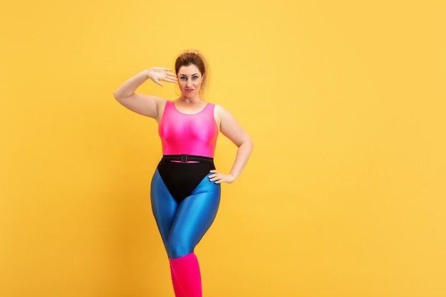 Young caucasian plus size female model's training on yellow background. Copyspace. Concept of sport, healthy lifestyle, body positive, fashion, style. Stylish woman posing confident like superhero.