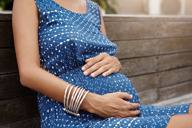 Young Caucasian pregnant woman wearing blue summer dress having rest on wooden bench during nice walk in urban park, touching her belly tenderly, feeling connected to her unborn baby. Cropped shot