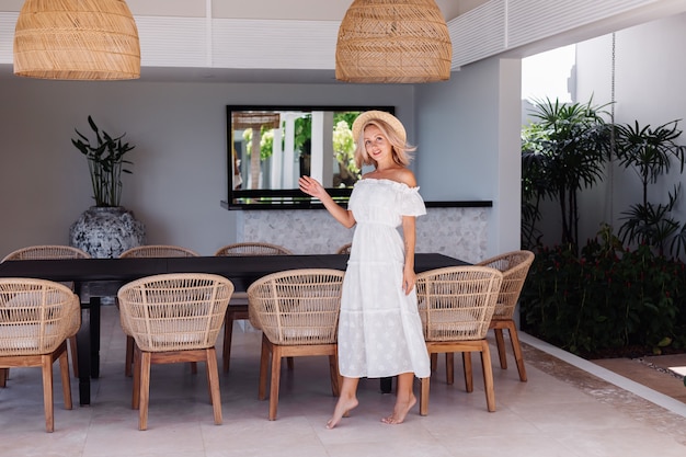 Foto gratuita donna giovane indoeuropea piuttosto bionda capelli in cucina all'aperto a villa donna con trucco leggero in abito bianco estivo e cappello di paglia in vacanza