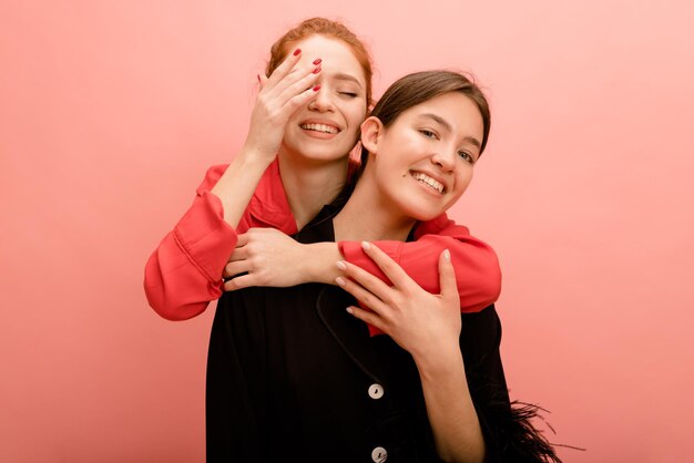 Young caucasian redhaired girl with closed eyes hugs brunette girlfriend from back on pink background Good relationship concept