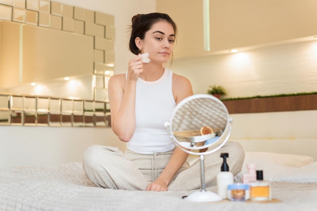 Free photo a young caucasian smiling woman is sitting on the bed and doing a facial massage