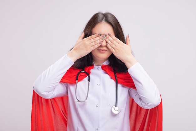 Giovane ragazza caucasica del supereroe che indossa l'uniforme del medico e lo stetoscopio che copre gli occhi con le mani