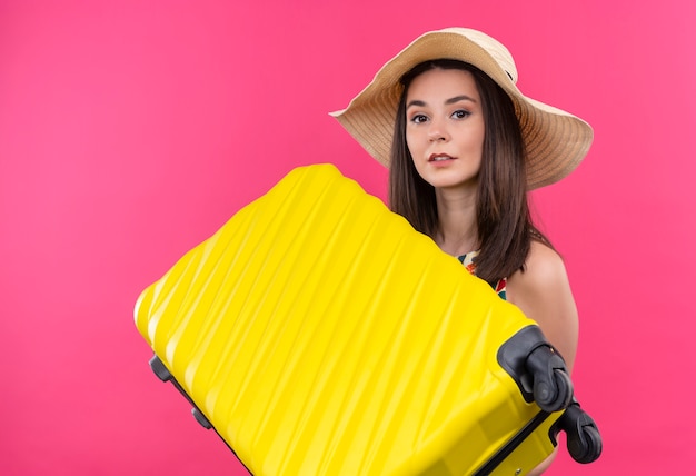Молодая кавказская девушка путешественника смотрит в камеру в шляпе, держа чемодан на изолированном розовом фоне