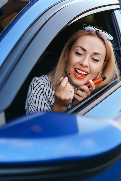 자동차 거울에 반사보고 립스틱을 적용하는 젊은 백인 여자.