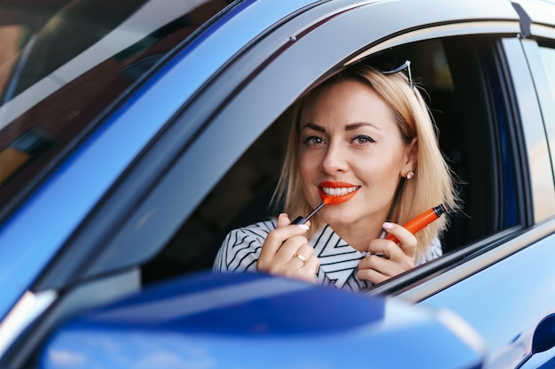 Young Caucasian woman applying lipstick looking at reflection in car mirror.