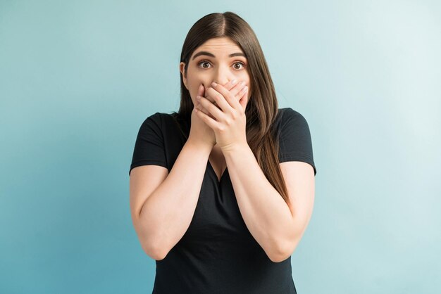 Young Caucasian woman covering mouth with hands in shock while making eye contact against plain background