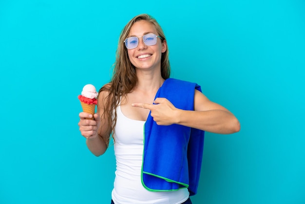 Молодая кавказская женщина держит мороженое и носит пляжное полотенце, изолированное на синем фоне и указывая на него