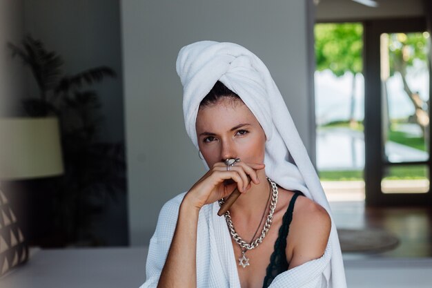 Young caucasian woman in hotel room in bathrobe and white towel on had with cigar.