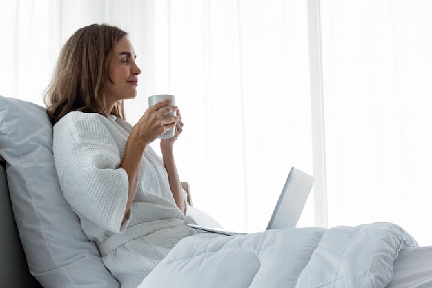 Молодая кавказская женщина в белой пижаме с белым держит чашку кофе и напитка и находит информацию с ноутбука на кровати в спальне дома. женщина пьет кофе и работает на ноутбуке.