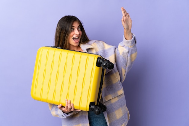 Молодая кавказская женщина изолирована на фиолетовой стене в отпуске с чемоданом и салютует