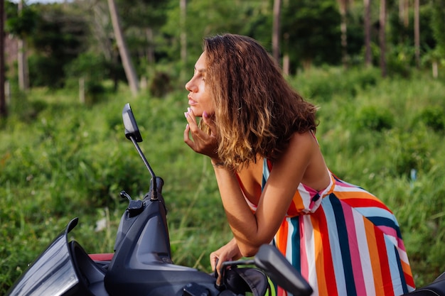 Giovane donna caucasica guarda nello specchio del motociclo sul campo tropicale