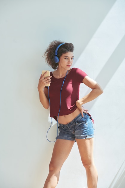 Free photo young caucasian woman in shorts, with headphones and smartphone, posing against white wall