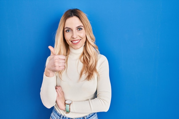 Young caucasian woman standing over blue background doing happy thumbs up gesture with hand. approving expression looking at the camera showing success.