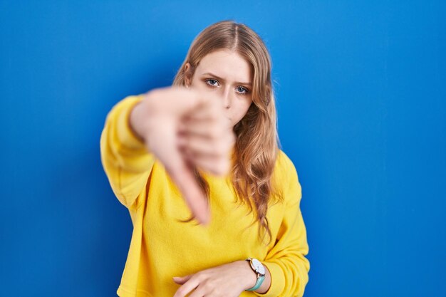 青い背景の上に立つ若い白人女性は不幸で怒っているように見え、拒絶と否定的で親指を下げてジェスチャーが悪い表情を示している