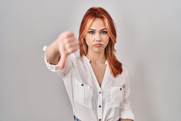 Бесплатное фото Молодая кавказская женщина, стоящая на изолированном фоне, выглядит несчастной и злой, показывая неприятие и негатив с опущенными большими пальцами и плохим выражением лица