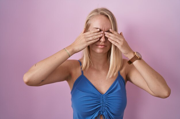 ピンクの背景の上に立っている若い白人女性は、疲労と頭痛、眠くて疲れた表情で目をこすります。視力の問題