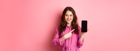 Young caucasian woman in stylish dress pointing finger at smartphone screen and smiling showing prom