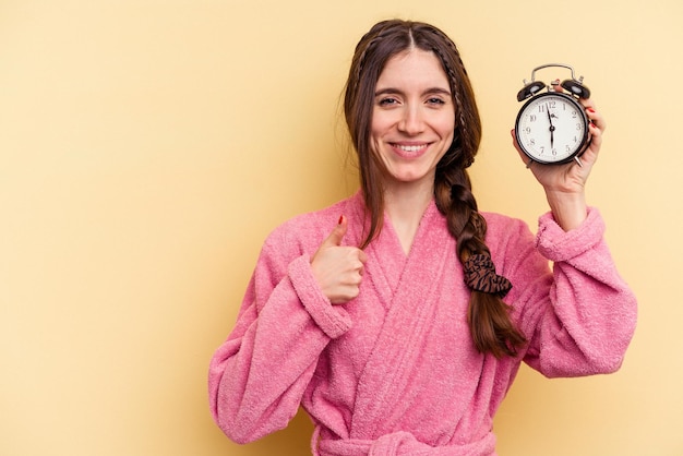 Young caucasian woman wearing a bathrobe holding a alarm clock isolated on yellow background smiling and raising thumb up