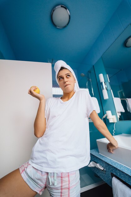 Young caucasian woman wearing towel on head and t-shirt in bathroom, hold apple.