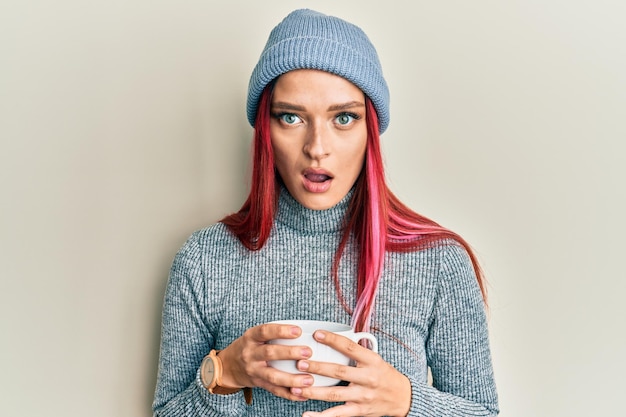 Young caucasian woman wearing winter cap and drinking a cup of hot coffee in shock face looking skeptical and sarcastic surprised with open mouth