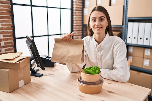 Young caucasian woman working at small business ecommerce holding take away food looking positive and happy standing and smiling with a confident smile showing teeth