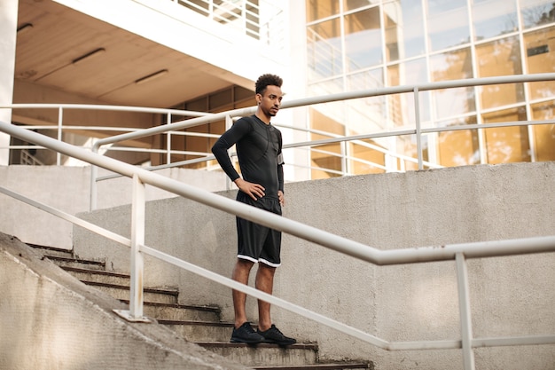 Young charming brunette dark-skinned man in shorts and black long-sleeved t-shirt going downstairs outside