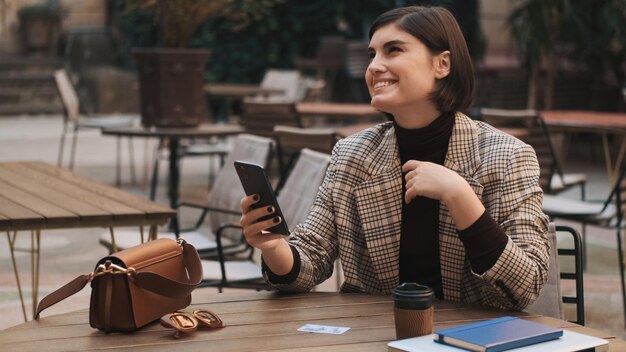 カフェの居心地の良い中庭でコーヒーブレイクでスマートフォンを使用して幸せそうに見える若い魅力的な実業家