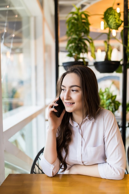 Young charming woman calling with cell telephone while sitting alone in coffee shop during free time, attractive female with cute smile having talking conversation with mobile phone while rest in cafe