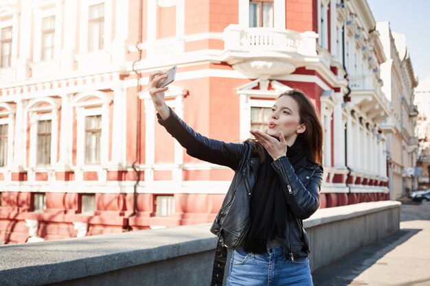 スマートフォンを保持している、外にいる間selfieを取って、街を歩いて、暖かい日を楽しんでいる若い魅力的な女性