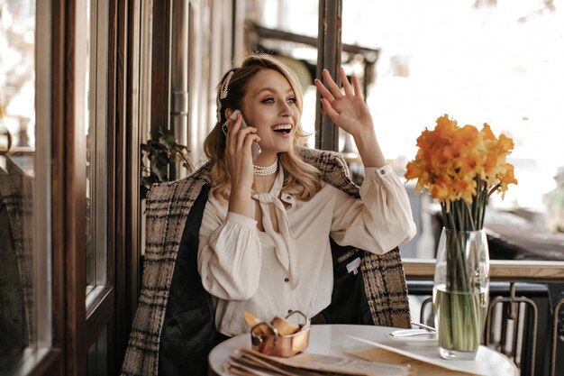 Young charming woman in trendy white blouse and checkered coat waves hand in greeting talks on phone and smiles in cafe