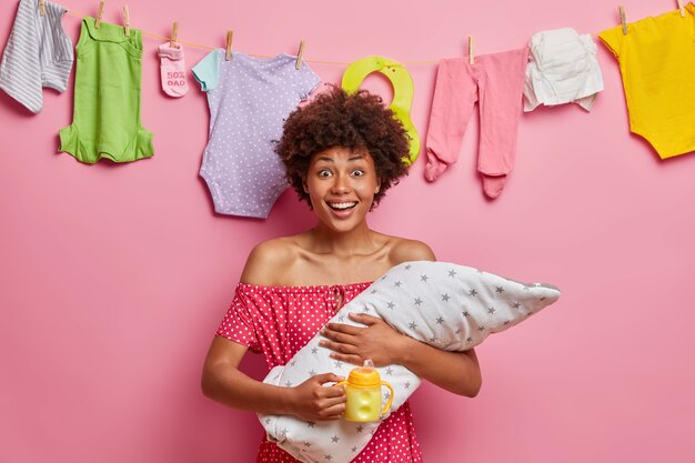 Young cheeful mother nursing baby, poses with newborn boy, feeds child, enjoys moterhood, spends time at home, cares of toddler, stands against pink wall with kids clothing hanging on rope