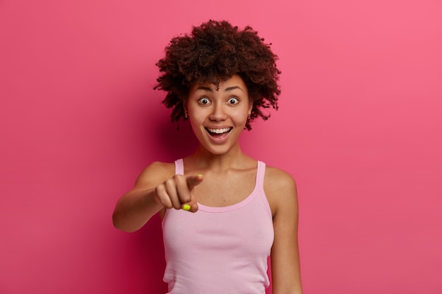 쾌활한 젊은 아프리카 계 미국인 여성이 앞쪽의 손가락을 가리키고, 믿을 수없는 것을 앞에서보고, 긍정적 인 것을 웃으며, 밝은 분홍색 벽 위에 고립 된 캐주얼 티셔츠를 입습니다.