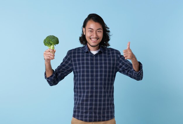 Young cheerful asian bearded man showing broccoli and hand thumbs up isolated on blue background