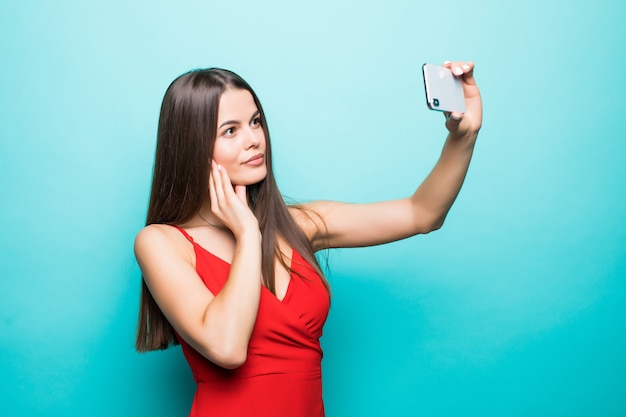 Young cheerful attractive brunette is smiling on the blue wall. She is taking selfie with her phone, wearing casual summer outfit and a hat