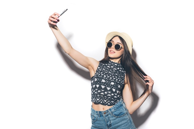 Young cheerful attractive brunette woman is smiling on the white wall taking selfie with her phone, wearing casual summer outfit and a hat