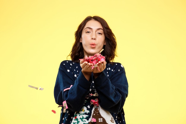 Young cheerful beautiful brunette girl in cosy knited sweater smiling with handful of confetti over yellow background.