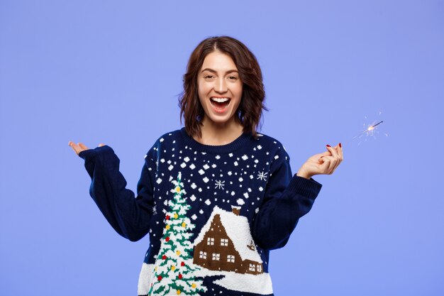 Young cheerful beautiful brunette girl in cosy knitted sweater smiling holding bengal lights over blue wall