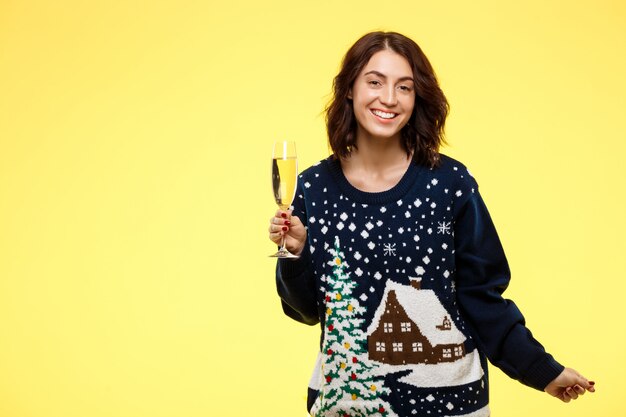 Young cheerful beautiful brunette girl in cosy knitted sweater smiling holding glass of champagne over yellow wall