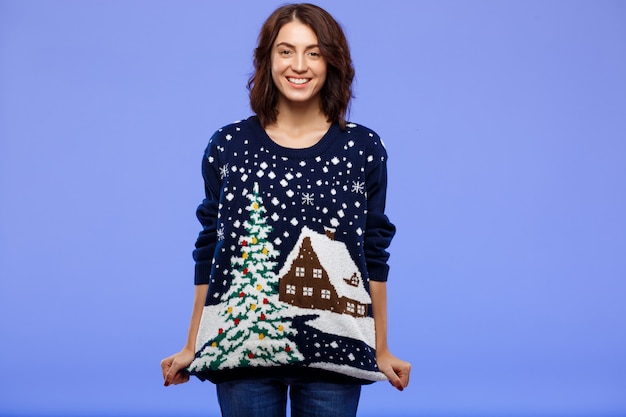 Free photo young cheerful beautiful brunette girl in cosy knitted sweater smiling posing over blue wall