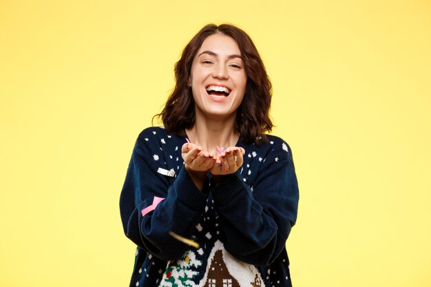 Young cheerful beautiful brunette girl in cosy knitted sweater smiling with handful of confetti over yellow wall