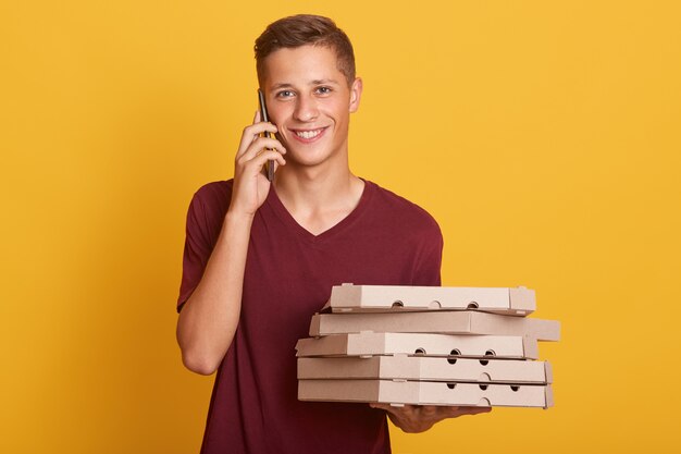 Young cheerful boy standing isolated on yellow in studio, holding cardboard boxes with pizza and smartphone in hands, having conversation, talking on phone, looking directly at camera.