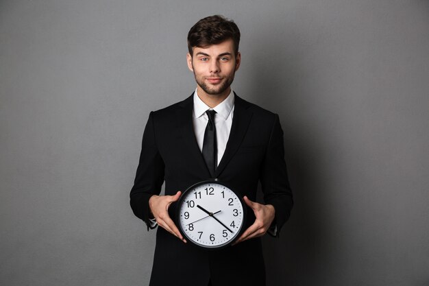 Young cheerful businessman in black suit holding big clock, 