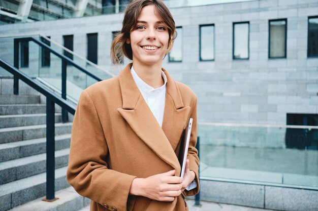 Young cheerful casual businesswoman in coat with laptop happily looking in camera outdoor