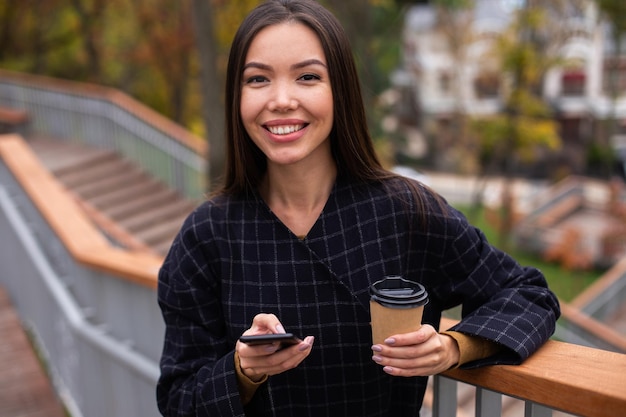 行くコーヒーと携帯電話でコートを着た若い陽気なカジュアルな女性は、都市公園のカメラで幸せそうに見ています