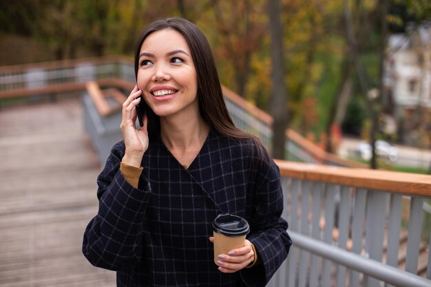 코트를 입은 젊고 쾌활한 캐주얼 여성, 커피와 함께 공원에서 휴대폰으로 행복하게 이야기하기
