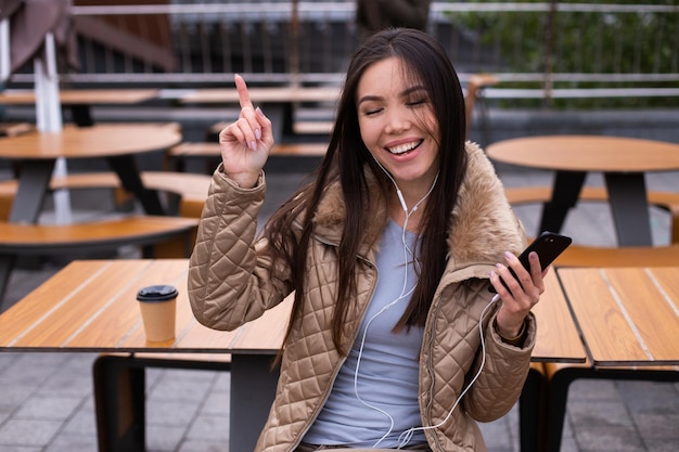 Young cheerful casual woman in down jacket happily dancing while listening music in earphones on cellphone outdoor