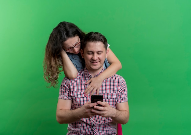 Young cheerful couple displeased woman standing behind her boyfriend and looking at his smartphone while he texting with somebody standing over green wall